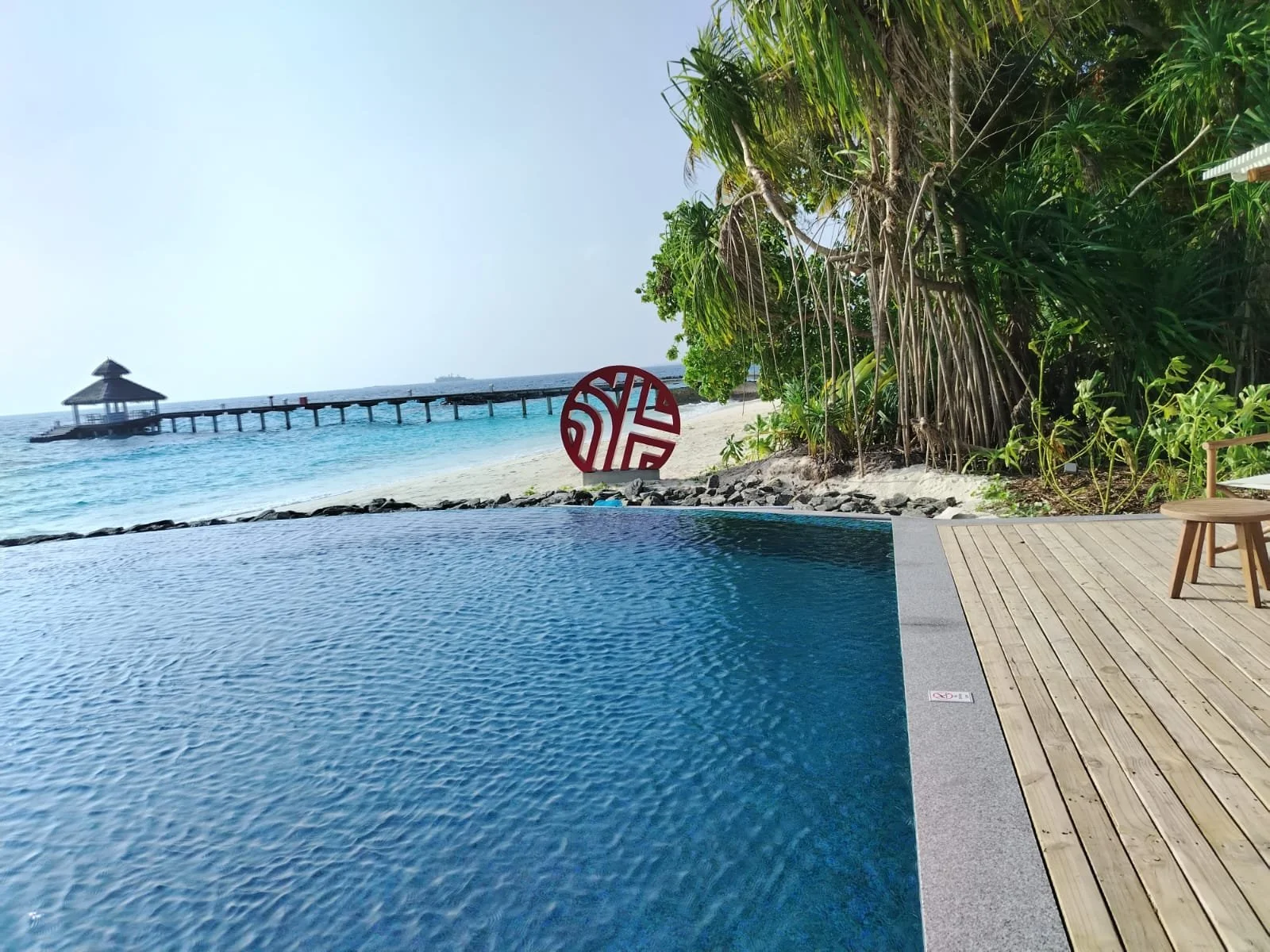 A luxurious infinity pool with a view of the beach and ocean, a pier with a gazebo extending into the water, lush greenery on the right side, and a decorative red globe sculpture on the sand.