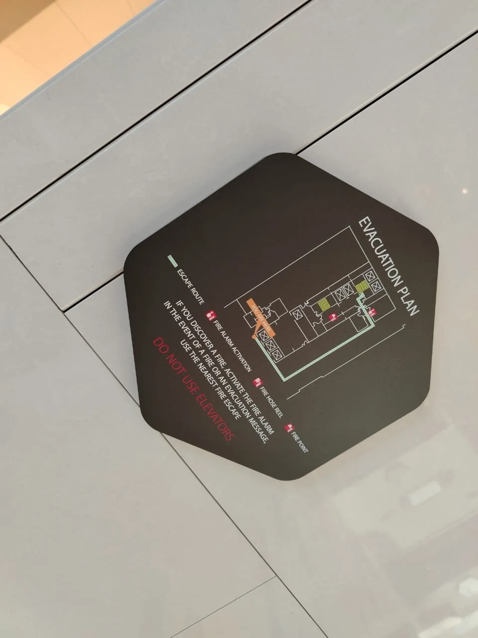 Evacuation plan sign on the floor showing escape route, fire alarm, fire hose reel, fire exit, fire front, and instructions to use nearest fire escape and not elevators.