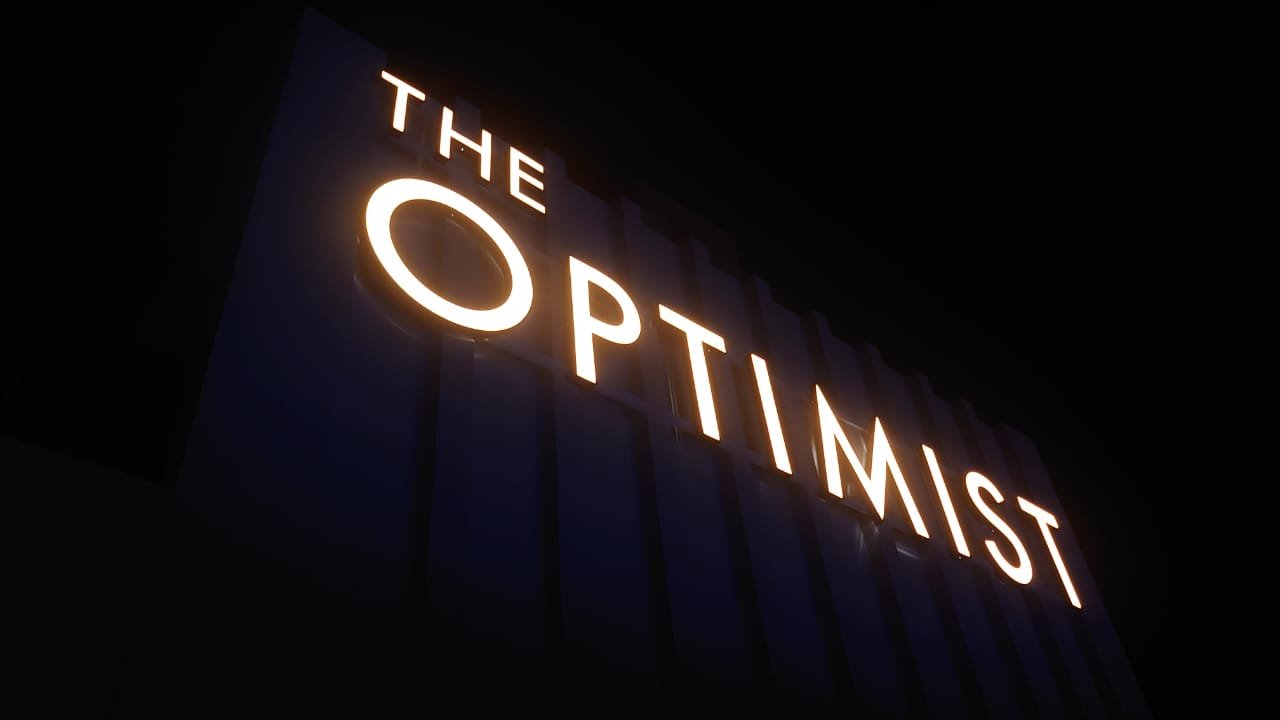 Illuminated sign reading "The Optimum" against a dark background.