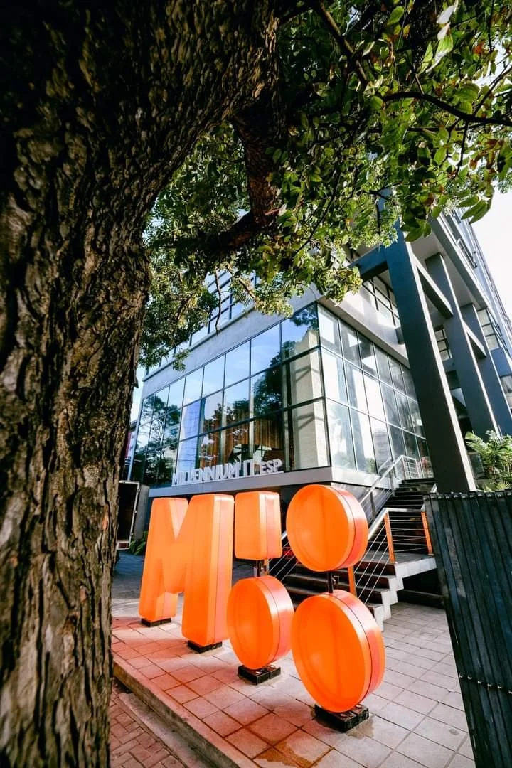 Modern building with large glass windows and orange decorative letters and circles outside, partially obscured by a tree in the foreground.