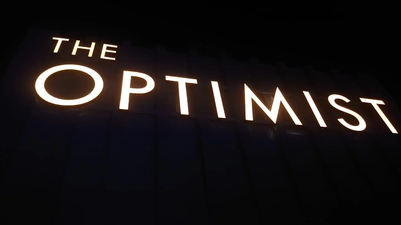 Illuminated sign displaying the words 'THE OPTIMIST' against a dark background.