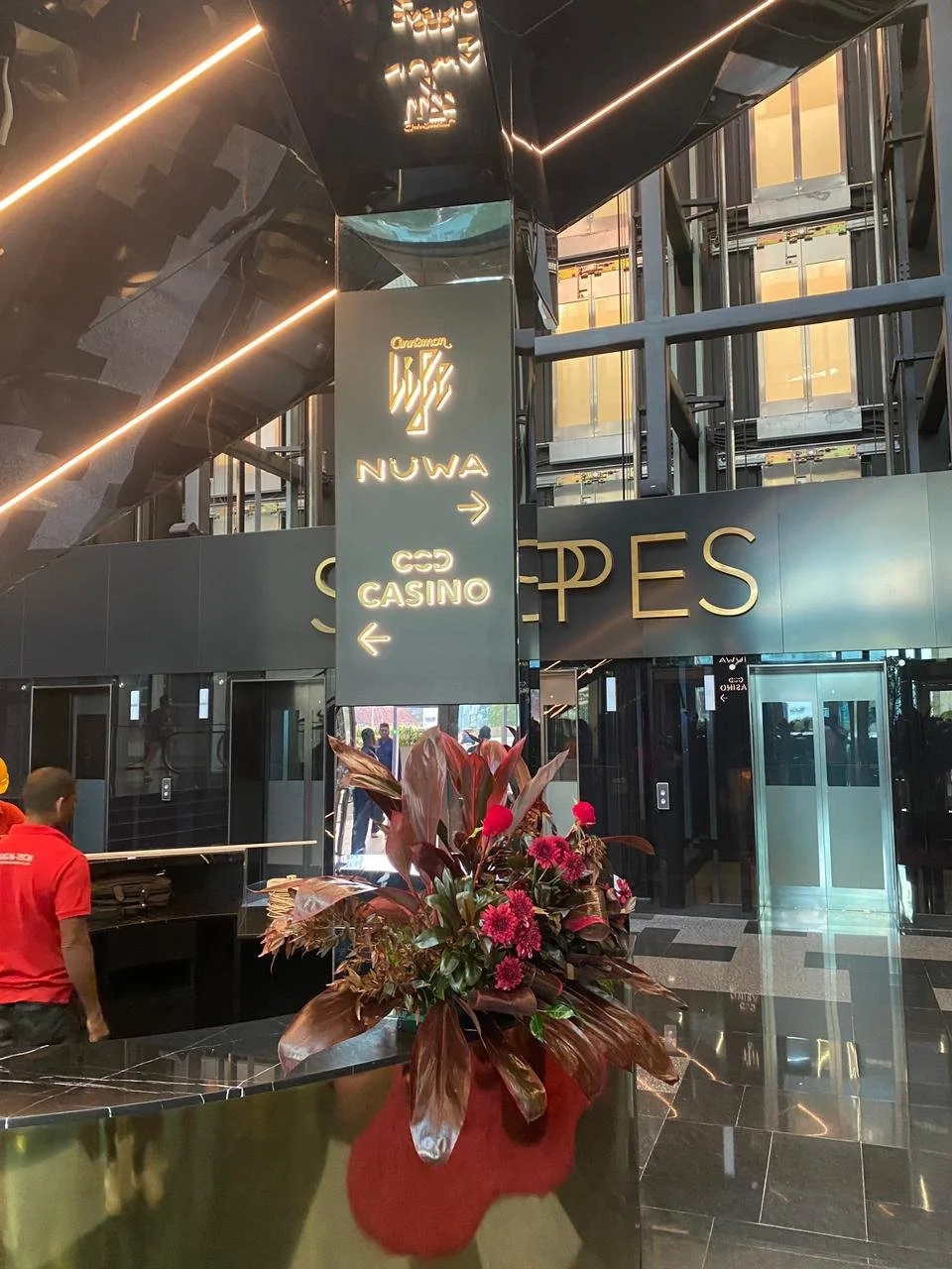 Interior view of a modern building lobby with black and gold decor, a sign indicating directions to NUWA and a casino, a flower arrangement with pink flowers and brown leaves on a black counter, and an elevator visible in the background.