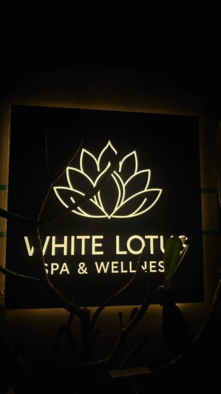 Illuminated sign for White Lotus Spa & Wellness featuring a lotus flower logo, partially obscured by plant branches in the foreground.