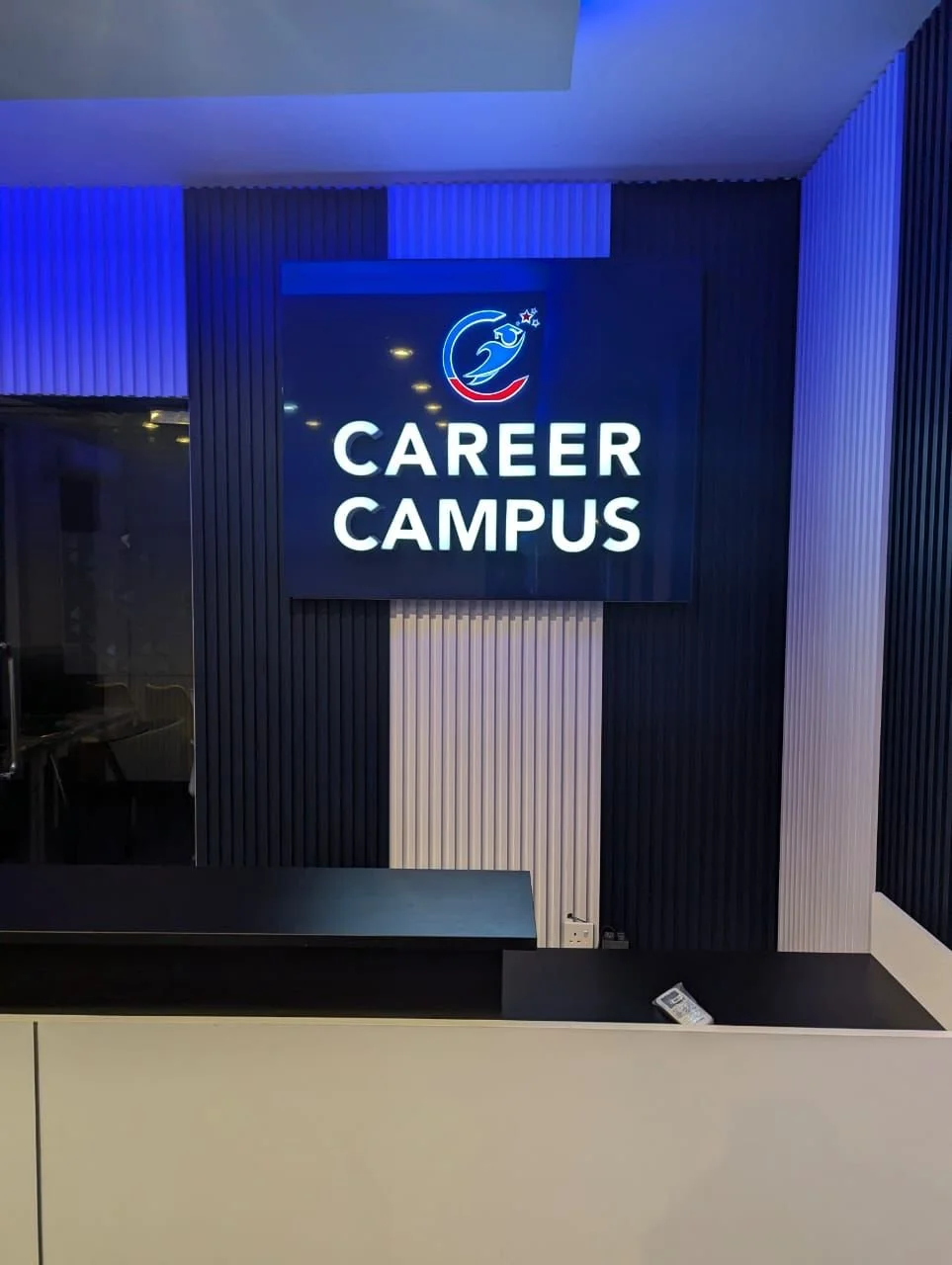 Neon sign that reads 'Career Campus' with a logo of a stylized bird and stars, mounted on a dark, ribbed wall.