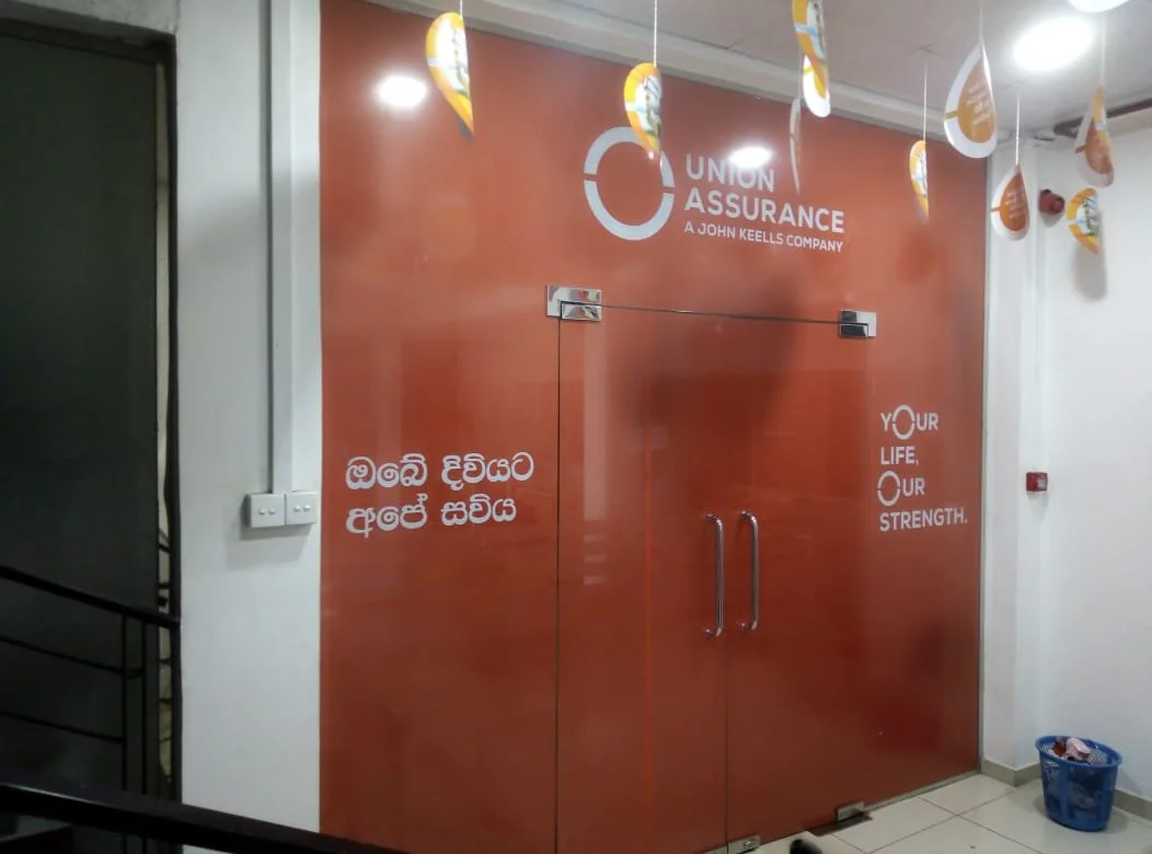 Entrance to Union Assurance office with a brown wooden door, white signage displaying the company logo, and slogans 'Your Life, Our Strength' in English and Sinhala. Decorative hanging flags are above, and a small trash bin is to the right.