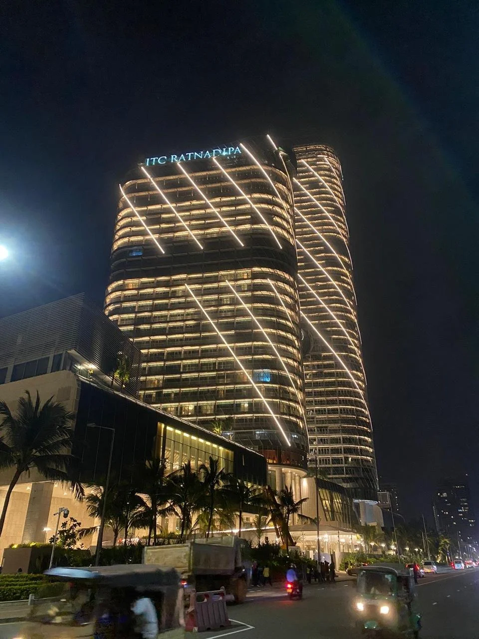Night view of a tall illuminated modern building with curved architecture and horizontal light lines, with the sign 'ITC RATTANP HOLDING' at the top, in an urban area with palm trees and vehicles on the street.