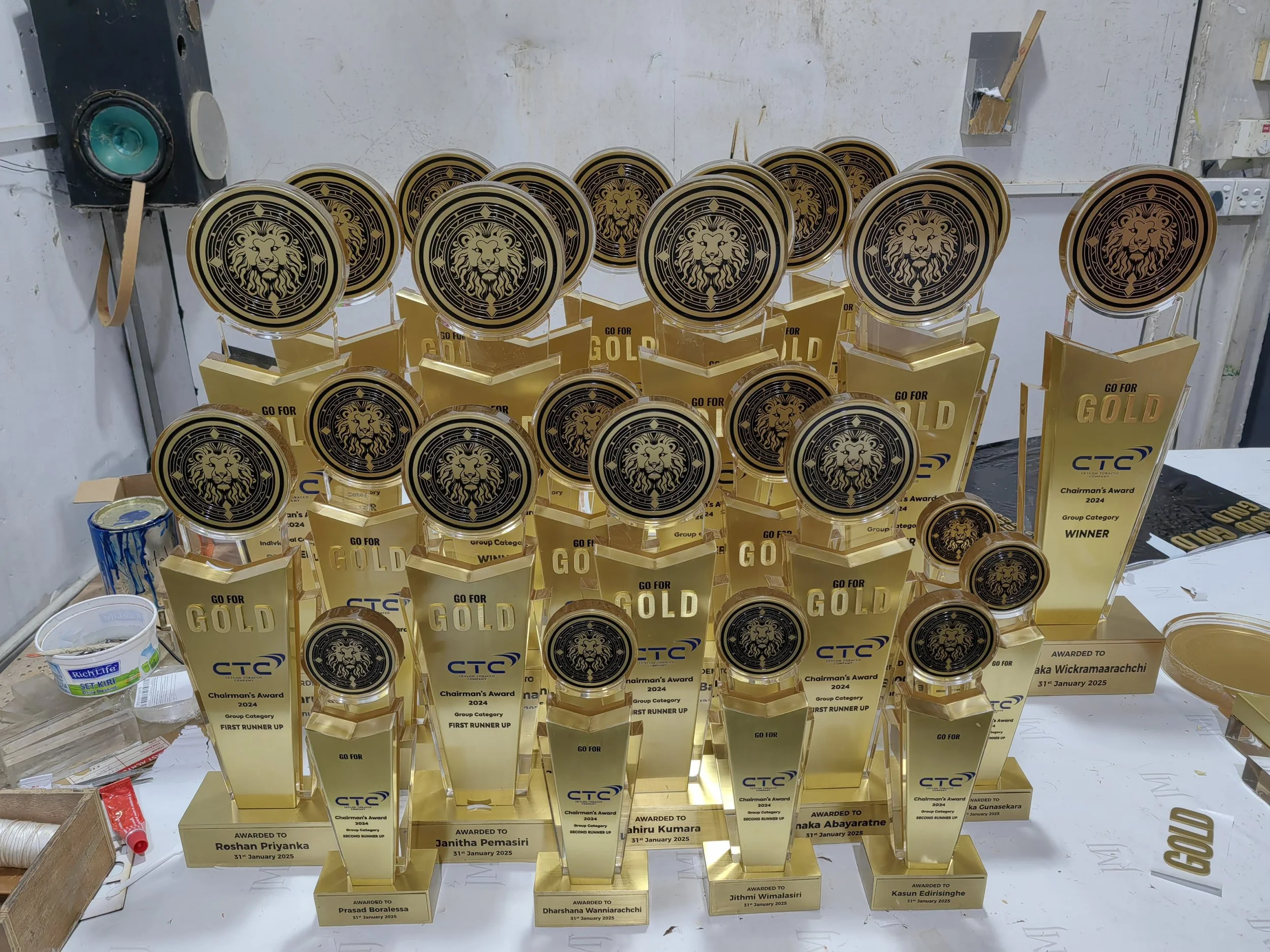 Several gold trophies with a lion's head design and inscriptions, displayed on a table against a white wall, with some paint cans and miscellaneous items nearby.