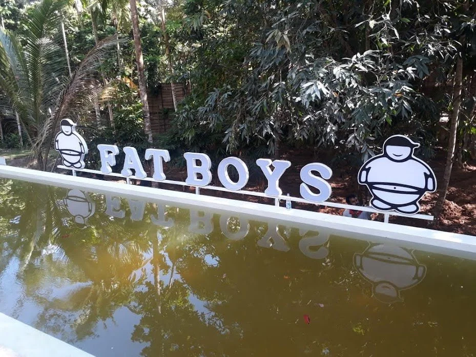 A sign with the words "Fat Boys" and cartoon illustrations of a chubby person on each side, located outdoors with trees and bushes in the background, reflected in a body of water.