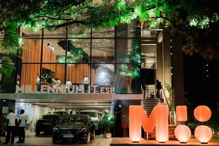 Outside view of a modern building at night with large glass windows and illuminated green foliage on top. People are visible inside, and there are luxury cars parked below. Large illuminated letters spelling 'M' and 'O' are in front of the building.
