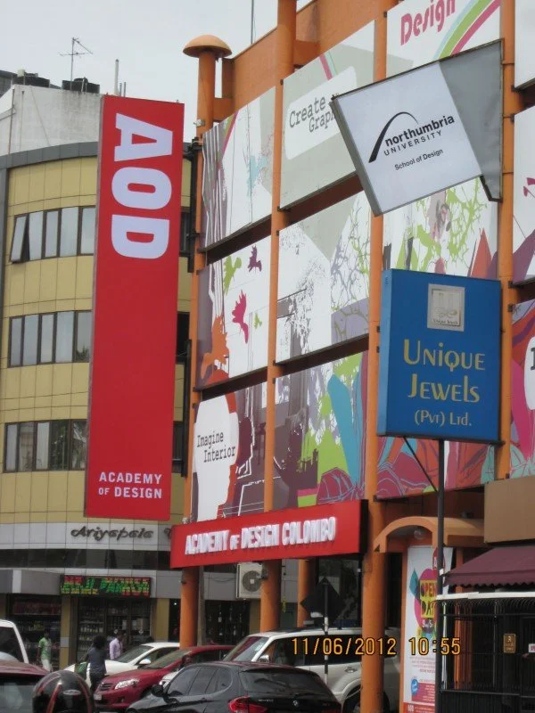 Outdoor building with multiple signs, including a red vertical sign for the Academy of Design, a Northumbria University sign, a Unique Jewels sign, and a banner for a design academy, with parked cars and people on the street.