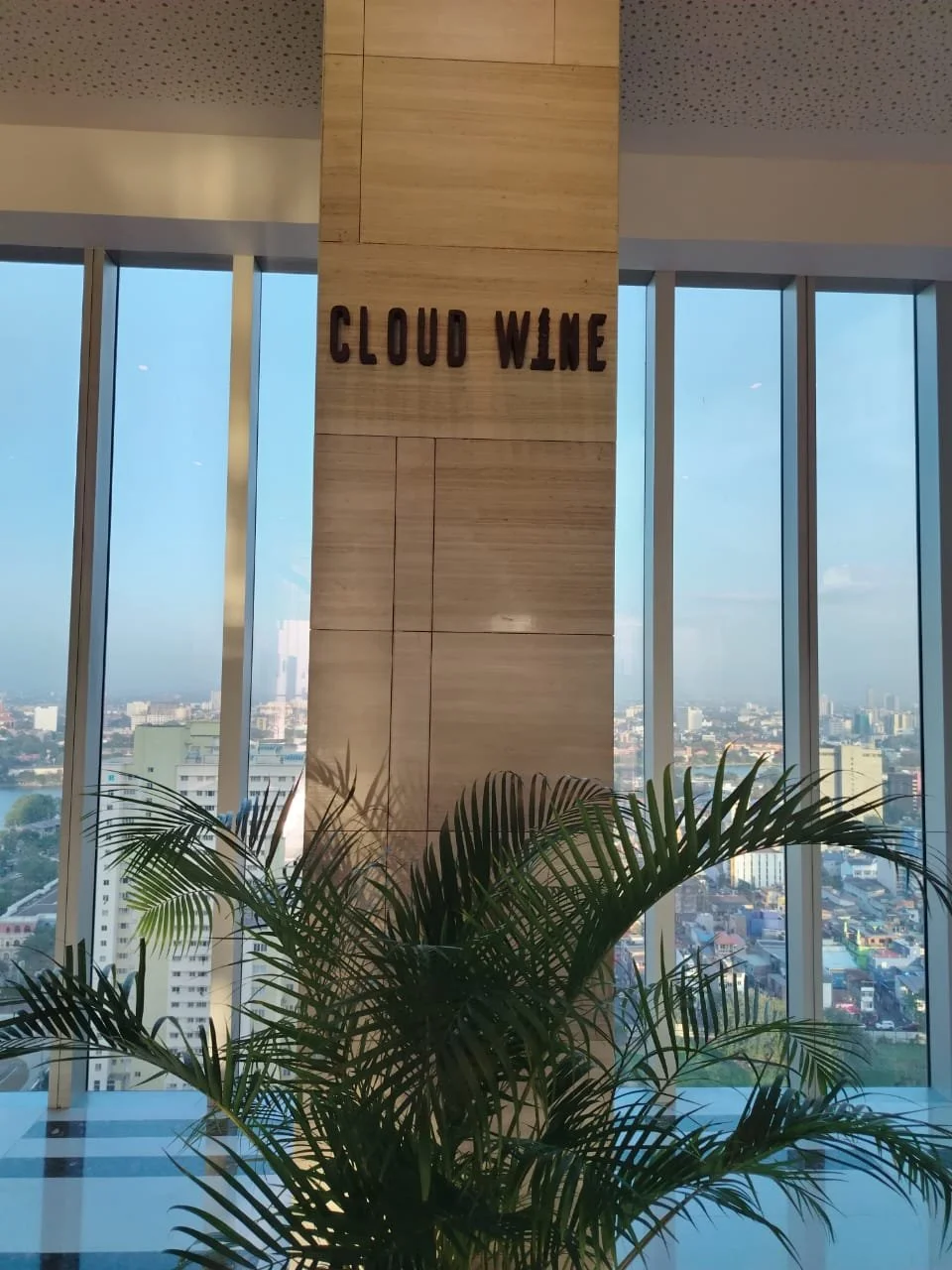 Interior view of a high-rise building with large floor-to-ceiling windows showing a cityscape outside. In the center, there is a beige marble pillar with the words 'CLOUD WINE' in dark letters. A green potted plant is in the foreground.
