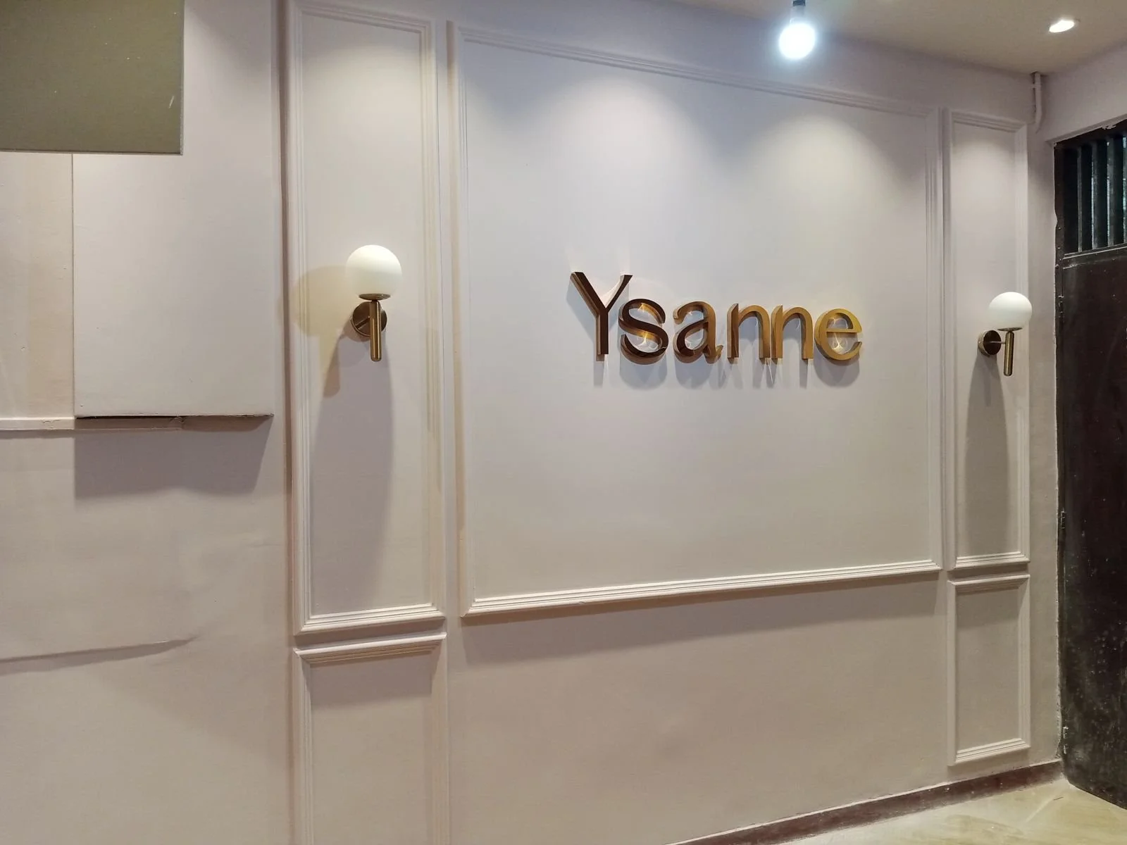 Wall with decorative panels, two wall sconces, and the word 'Ysame' displayed in gold-colored letters.