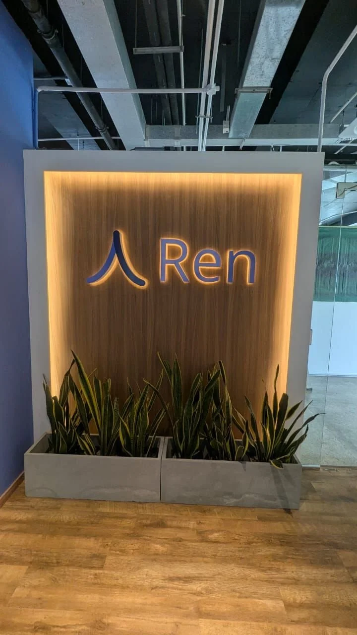 A decorative wall display at an office or building lobby with the company name 'Ren' and a Chinese character, illuminated with backlighting, with potted plants at the base on a wooden floor.