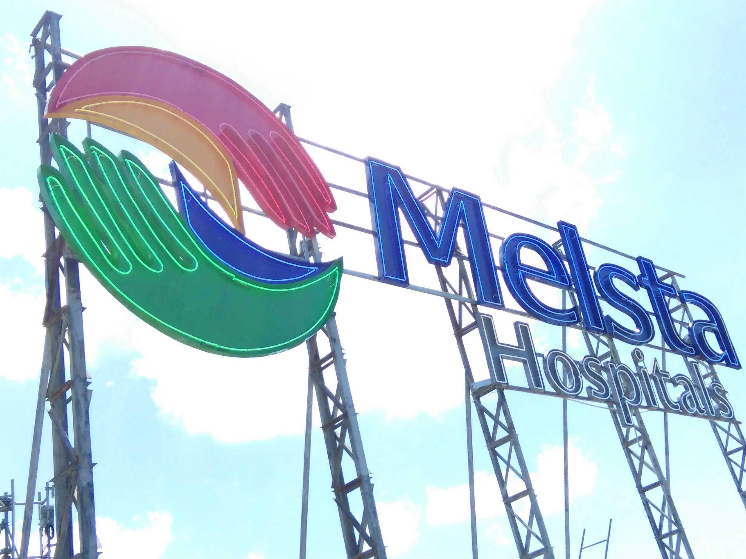 Colorful neon sign with the word 'METS' and a rainbow pattern against a blue sky with some white clouds.
