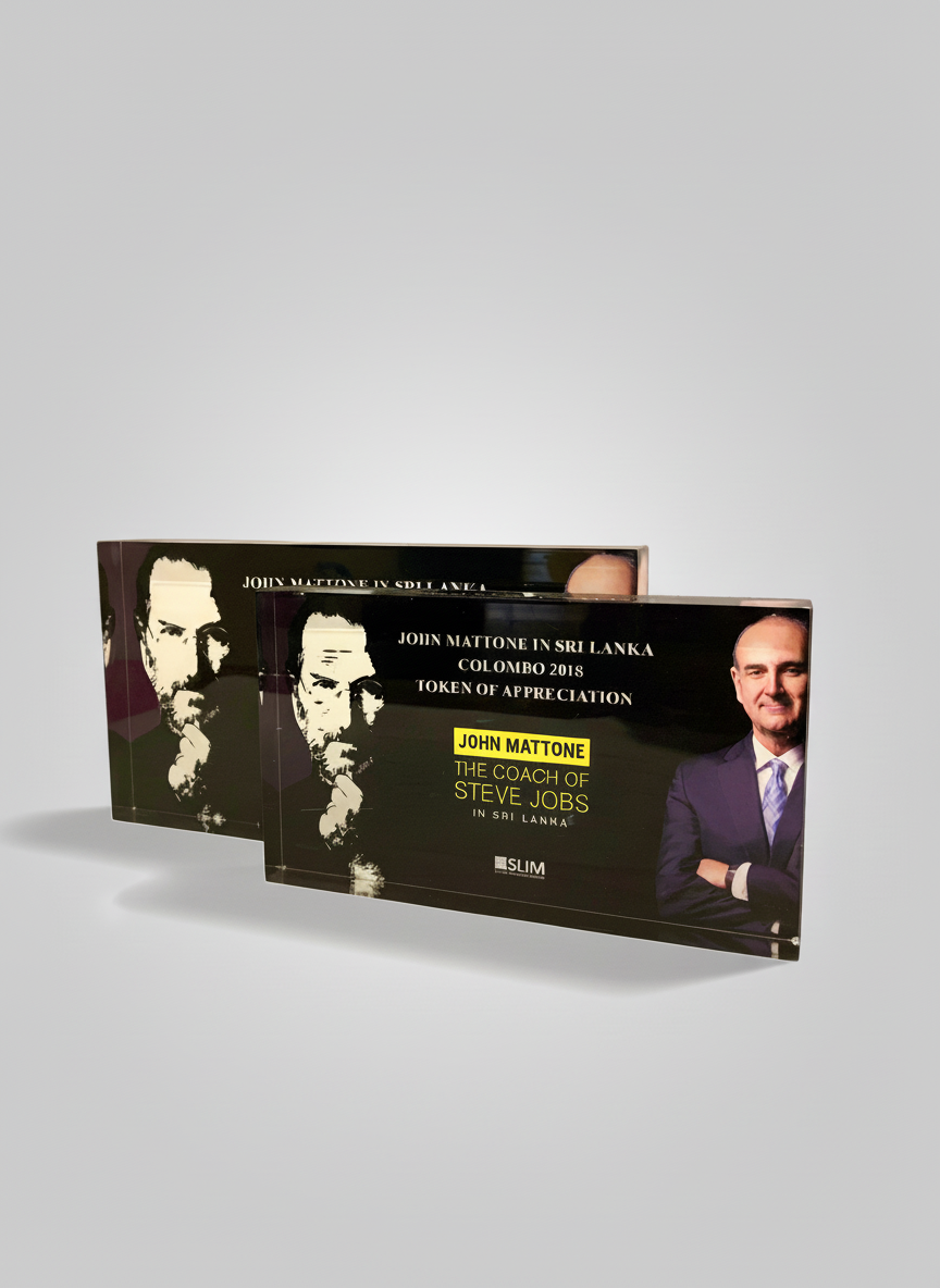 Two black plaques featuring a portrait of a man with a monocle and a hand on his chin, and a photo of a smiling man in a suit with arms crossed. The plaques include text about an event in Sri Lanka, Colombo 2018, with a mention of John Mattone and St