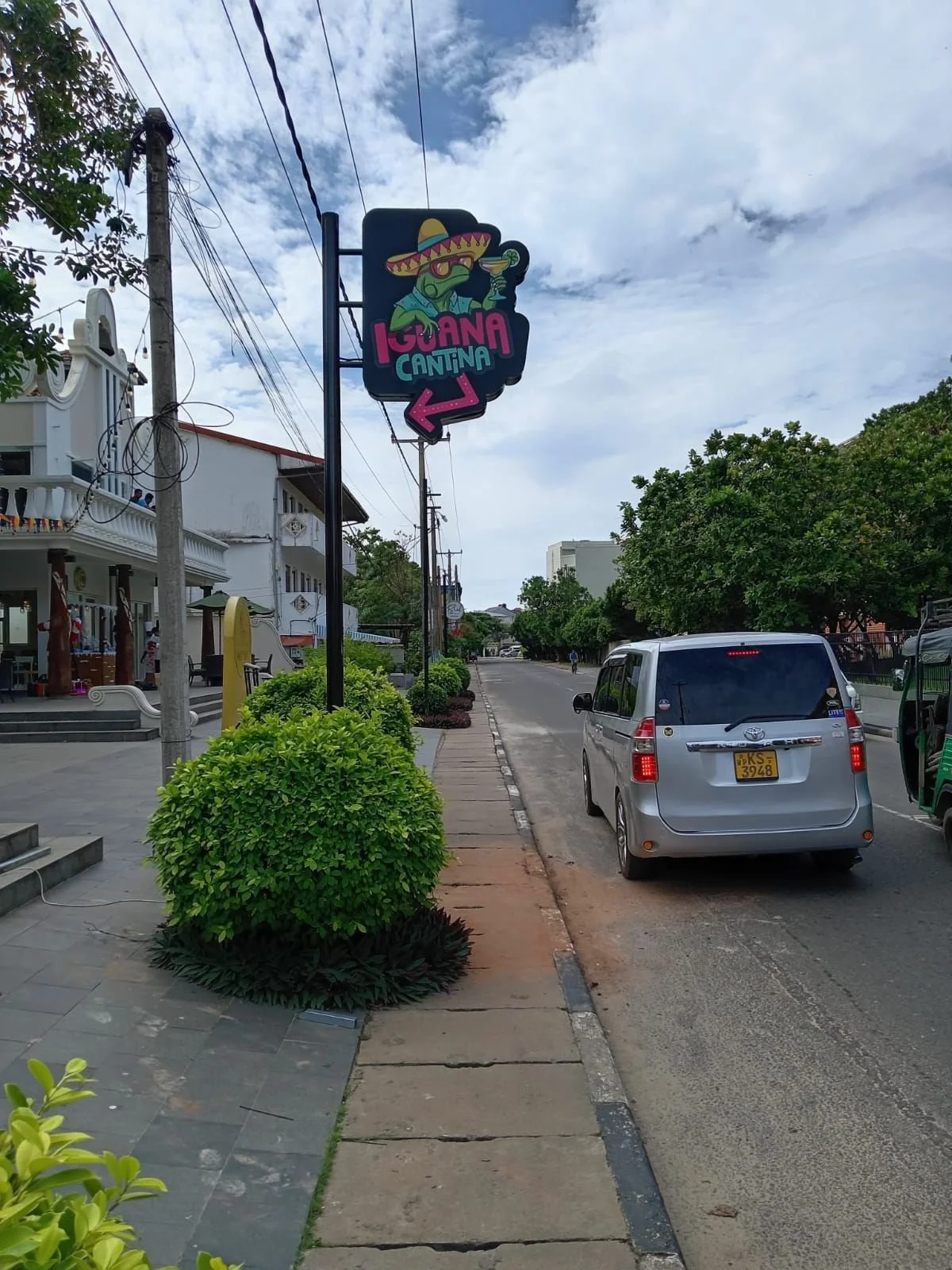 Street view with a sign featuring a frog wearing a sombrero and sunglasses, holding a cocktail, with the words 'IGUANA CANTINA' and an arrow pointing left. There is a white building with outdoor stairs and a porch on the left, green bushes along the 