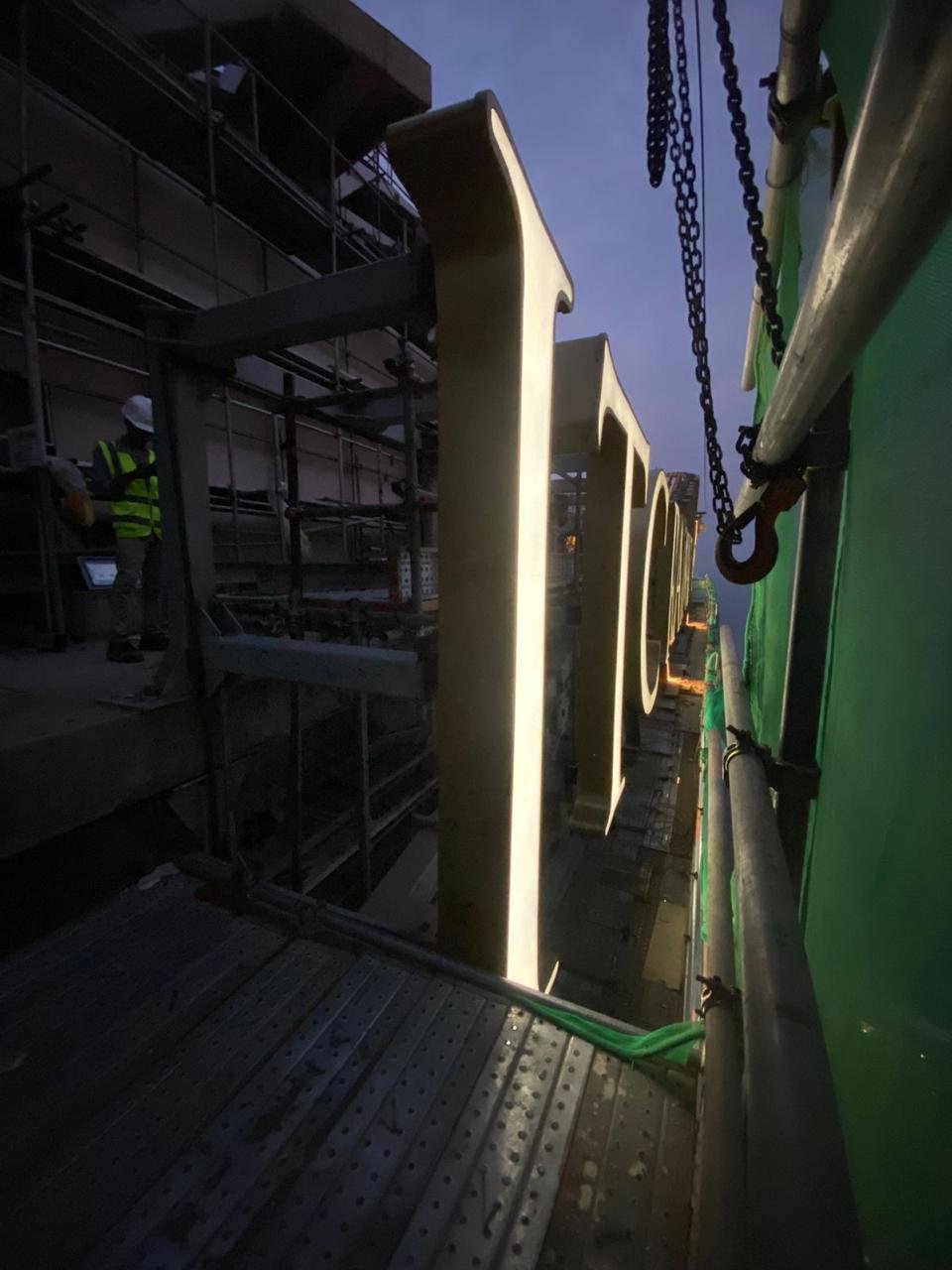 Close-up of large illuminated letters on a sign at dusk, with scaffolding and a construction worker in safety gear nearby.