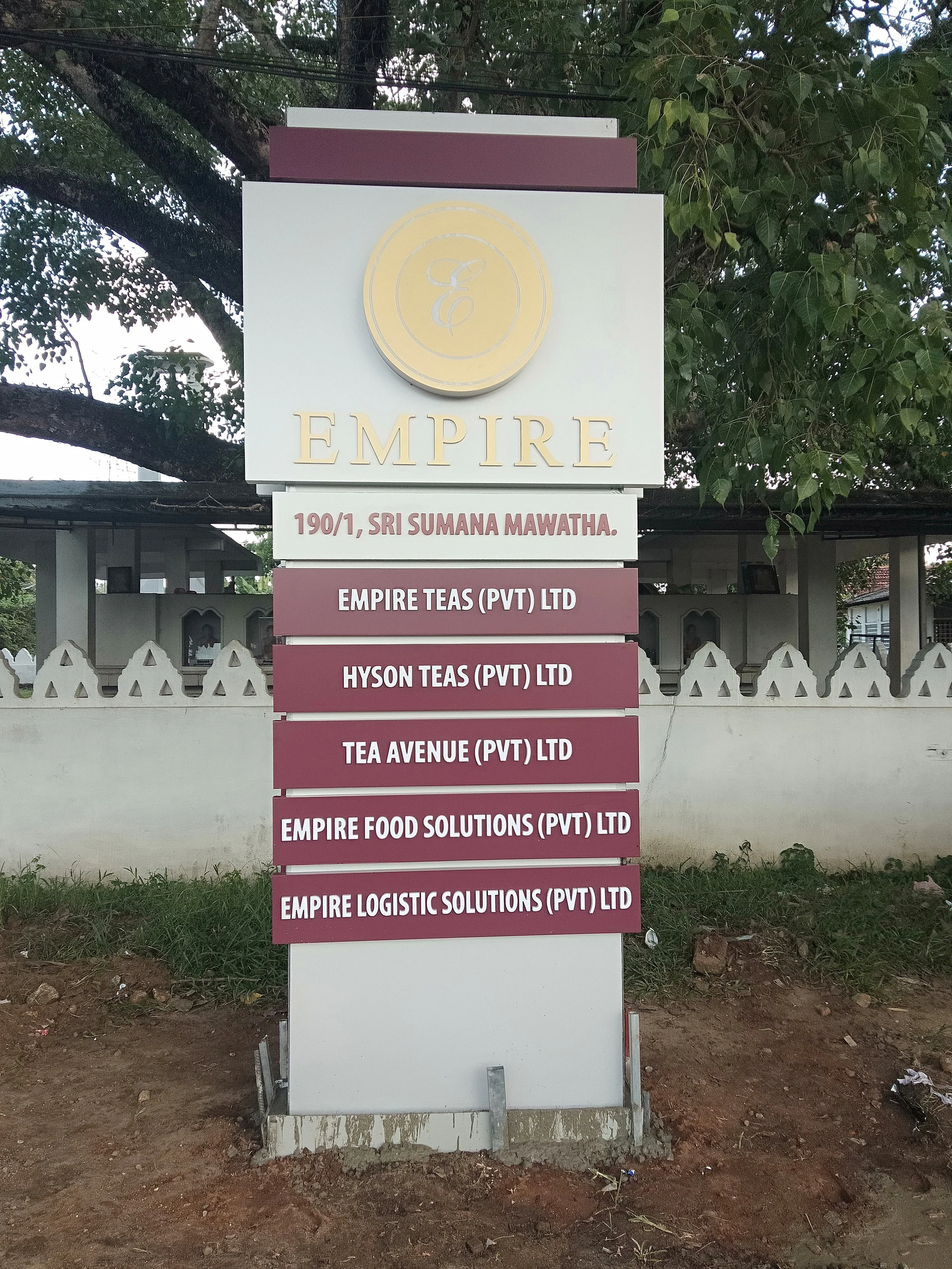 Signboard with multiple business names including Empire, Empire Teas, Hyson Teas, Tea Avenue, Empire Food Solutions, and Empire Logistic Solutions, located in front of a fence with trees in the background.