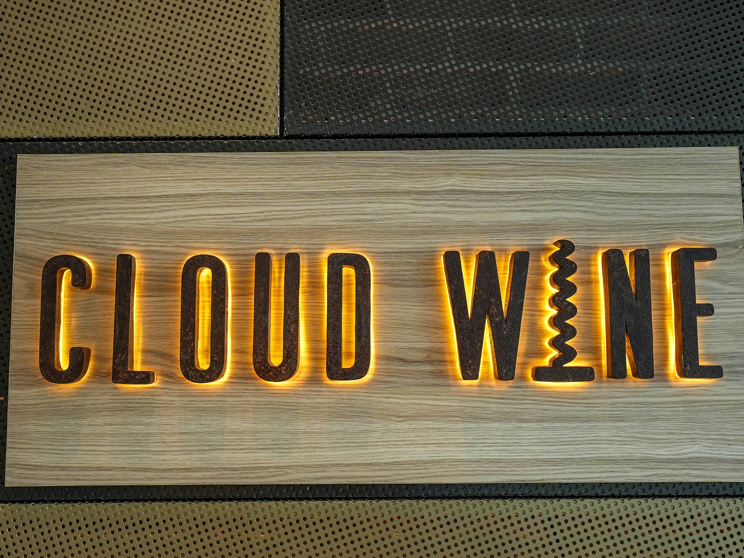 Close-up of a wooden sign with illuminated cut-out letters spelling 'CLOUD' and part of another word, mounted on a textured surface with black, yellow, and gray sections.
