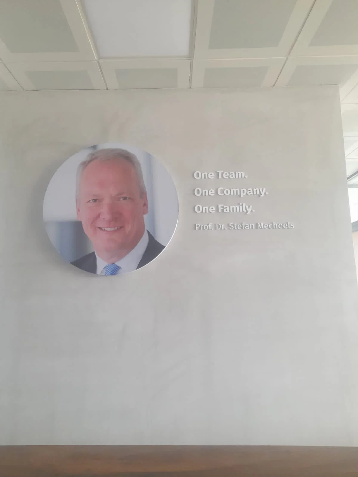 Poster on a wall featuring a professional headshot of a smiling man in a suit and tie, and the text: "One Team. One Company. One Family. Prof. Dr. Stefan Mechels".
