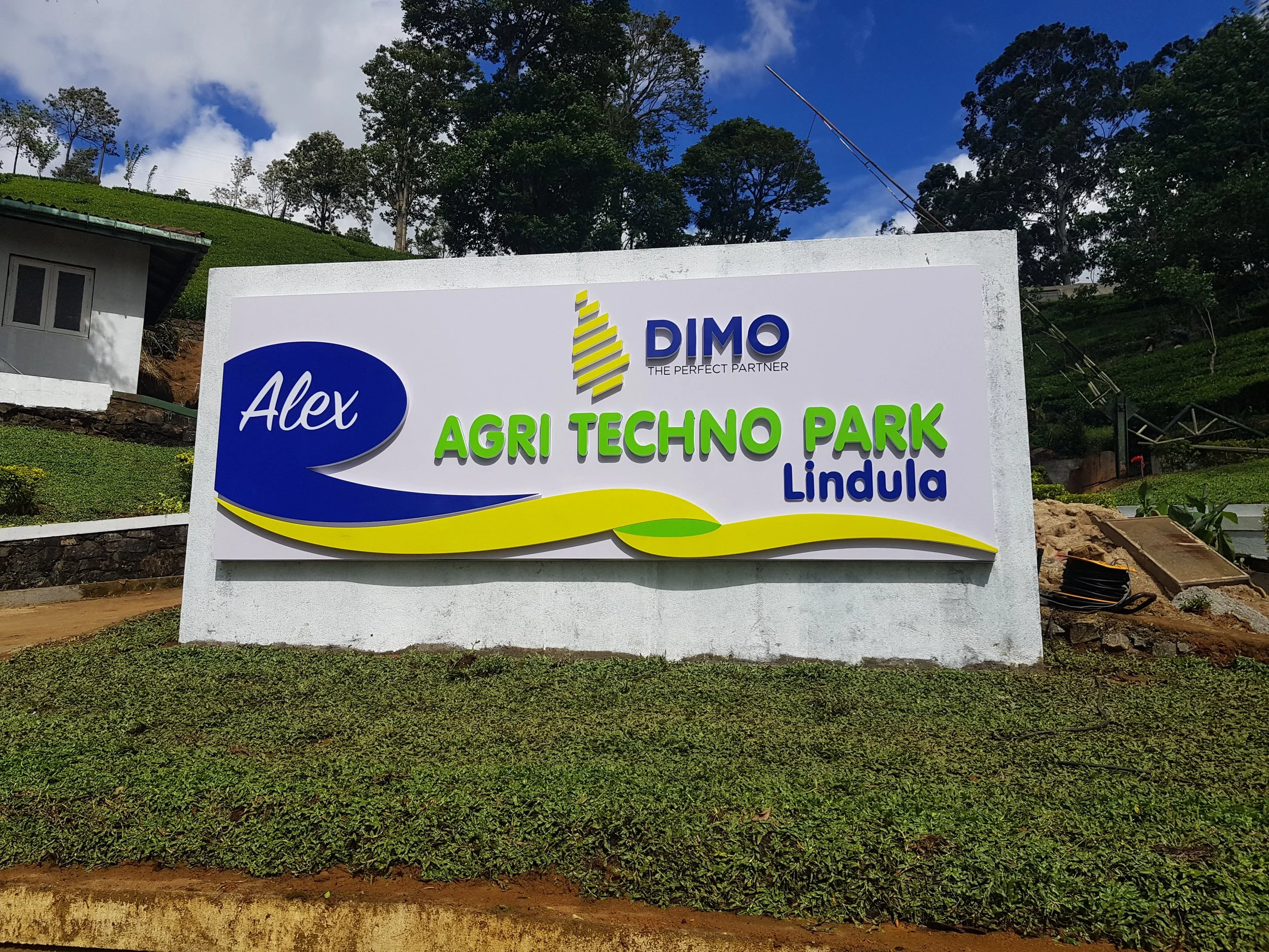 A sign at Agri Techno Park in Lindula with logos and text, under a partly cloudy sky, surrounded by green trees and grass.