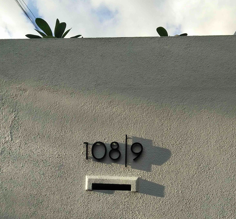 A white textured wall with black house numbers '108/9' and a mail slot beneath them, casting a shadow, against a background of a cloudy sky and green plant leaves.