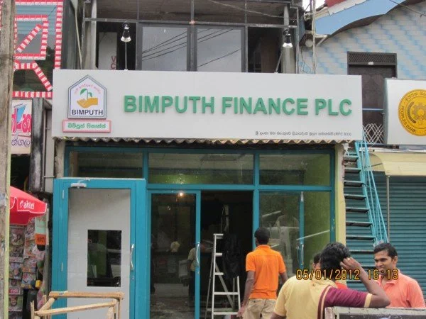 Outside the Bimpith Finance PLC office with a glass entrance, two men in orange and yellow shirts standing near the entrance, and a group of men sitting in front of the office.