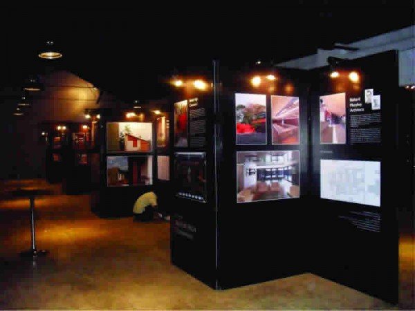 Exhibition with black display panels showcasing photographs and information, illuminated by warm ceiling lights, in a dark indoor space.