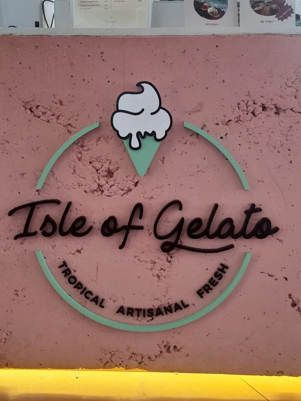 Sign for Isle of Gelato with a pastel pink background, a cartoon ice cream cone, and text indicating the shop specializes in tropical, artisanal, fresh gelato.