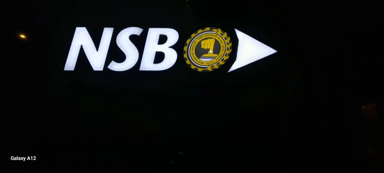 Illuminated sign with the abbreviation NSB, a logo with a yellow fist inside a black circle, and a right-pointing arrow, against a dark background.