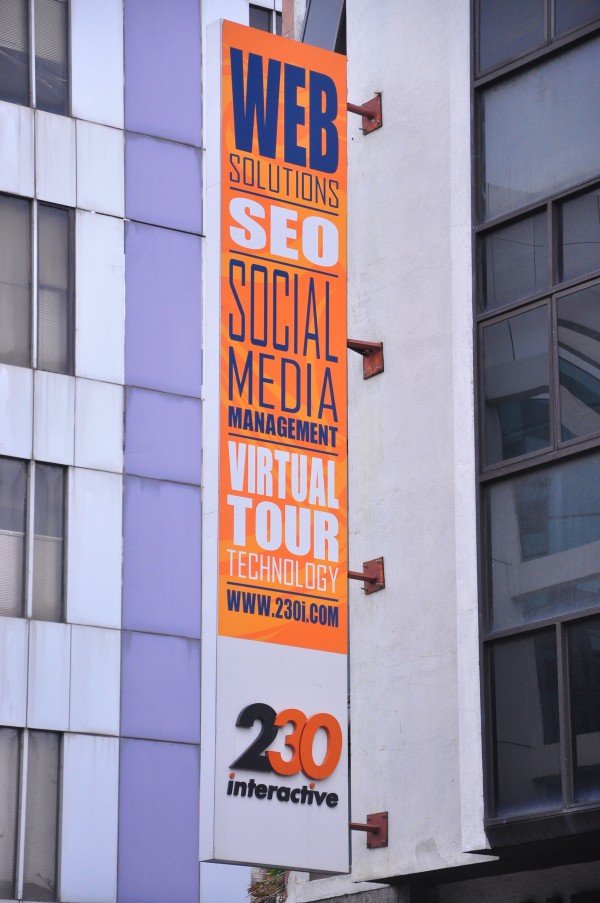 A vertical sign on a building advertising web solutions, SEO, social media, management, virtual tour technology, and a website URL, with a logo that reads '230 interactive' at the bottom.