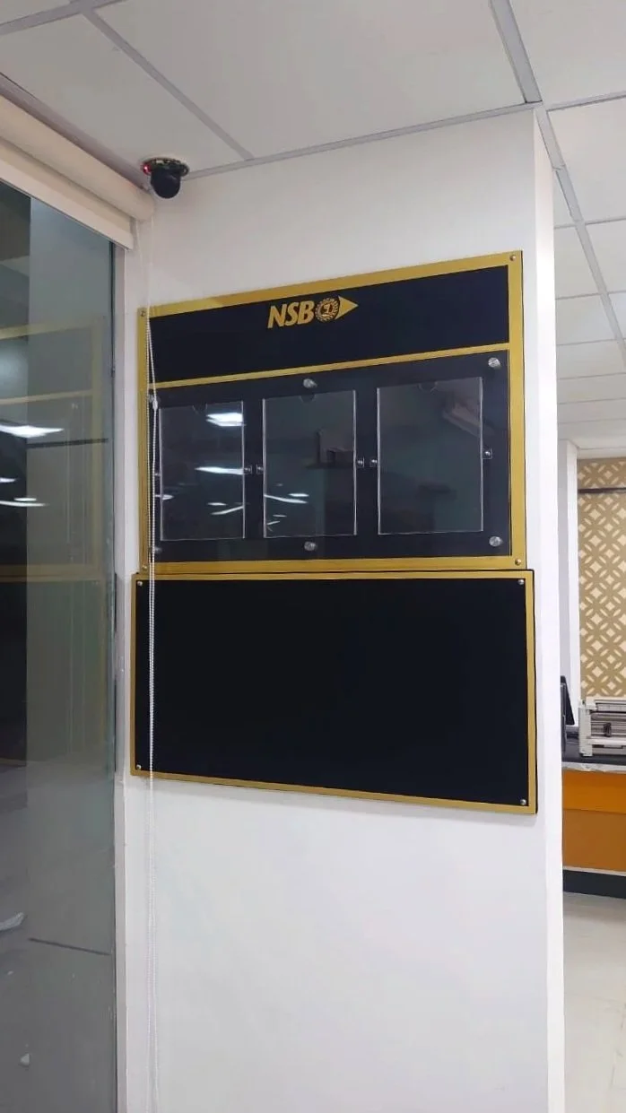 A black notice board with a gold trim and a glass display case on a white wall. The top left corner features a sign reading 'NSB' with an arrow pointing right.