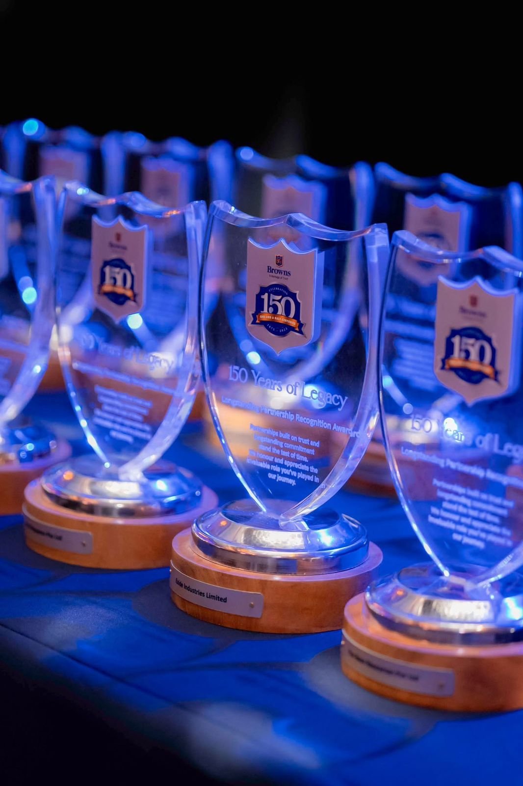 Glass awards with a shield shape, blue and orange logo honoring 150 years of legacy, placed on wooden bases, illuminated in blue light at a celebration event.