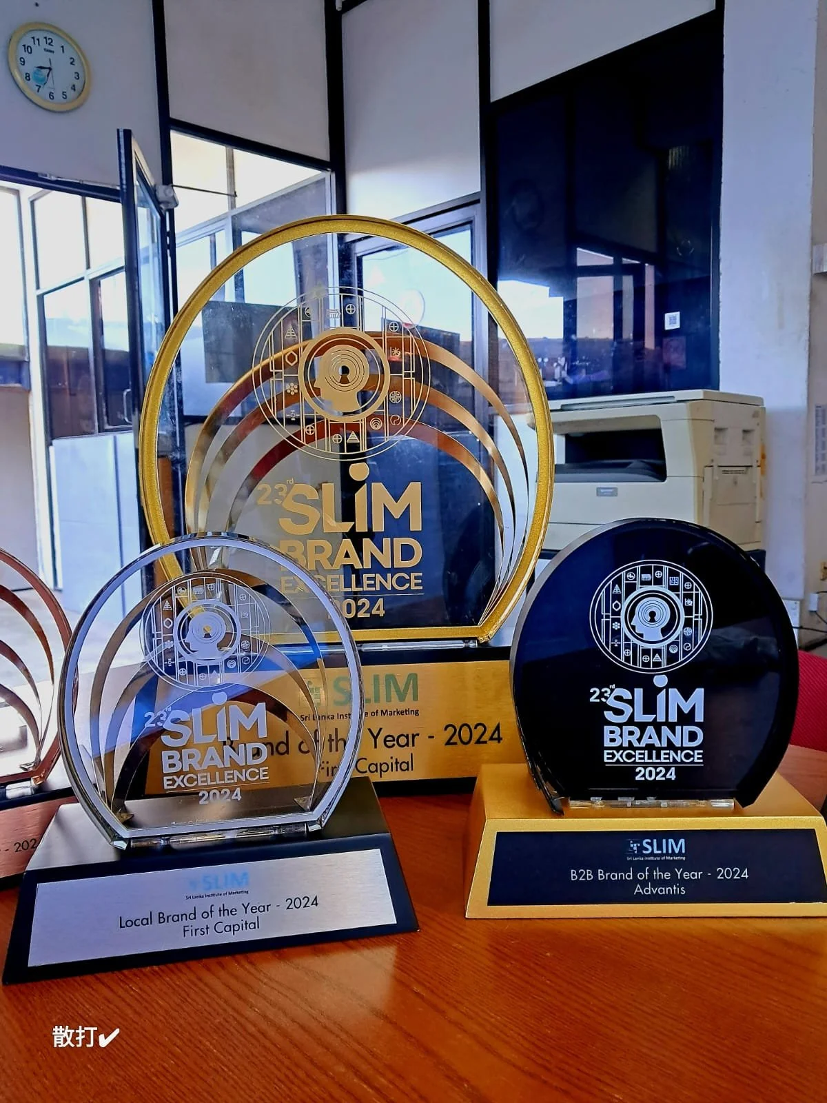 Several awards for the 23rd SLIM Brand Excellence 2024, including 'Local Brand of the Year - 2024 First Capital' and 'B2B Brand of the Year - 2024 Advantis,' displayed on a table with a clock on the wall in the background.