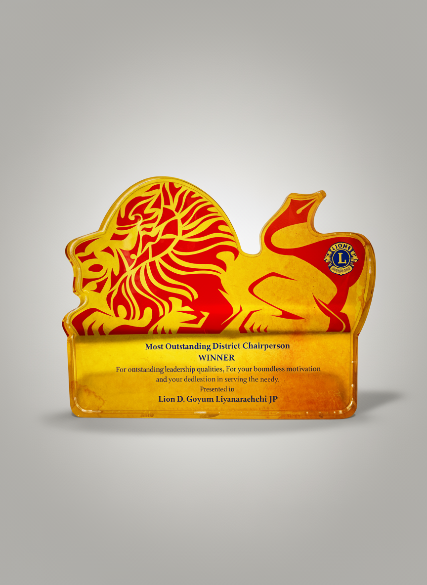 A trophy with a red and yellow lion graphic on top. The trophy has an inscription congratulating Lion D. Goyum Liyanaraehi JP for being the most outstanding district chairperson, with a message about leadership and service to the needy.