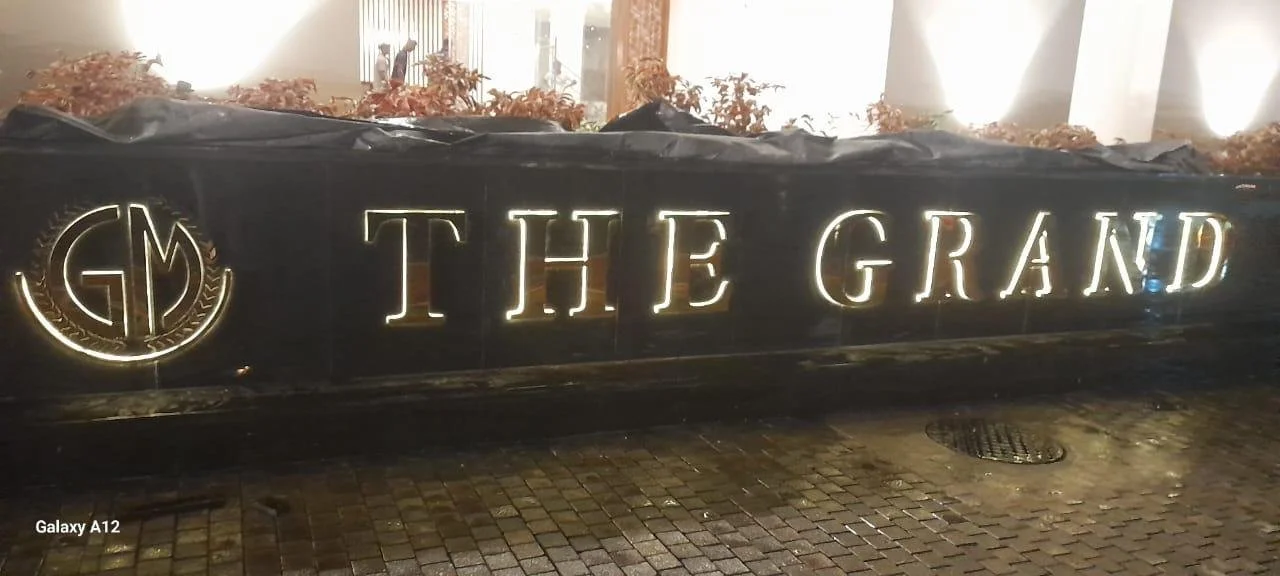 Illuminated sign reading 'THE GRAND' with a logo on the left, set against a dark background, outside at night with wet cobblestone pavement.