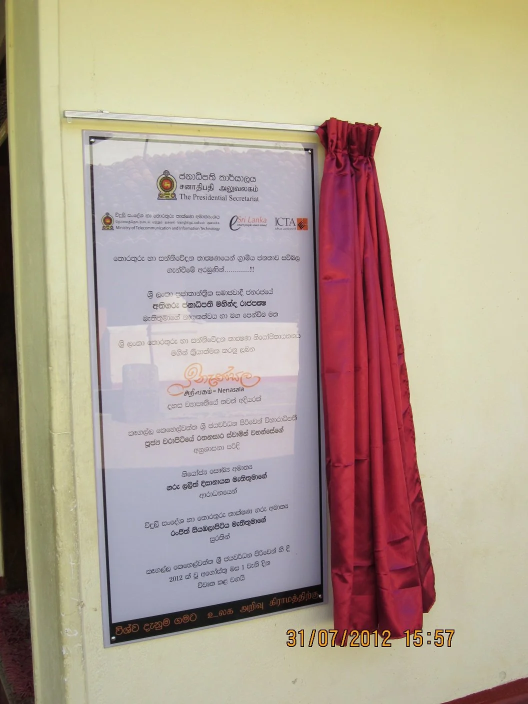 An unveiling ceremony for a sign with text in Sinhala and English, including logos of Sri Lanka's Presidential Secretariat, Ministry of Telecommunication and Information Technology, ICTA, and Nenasala, with a red curtain pulled to the side, indicatin