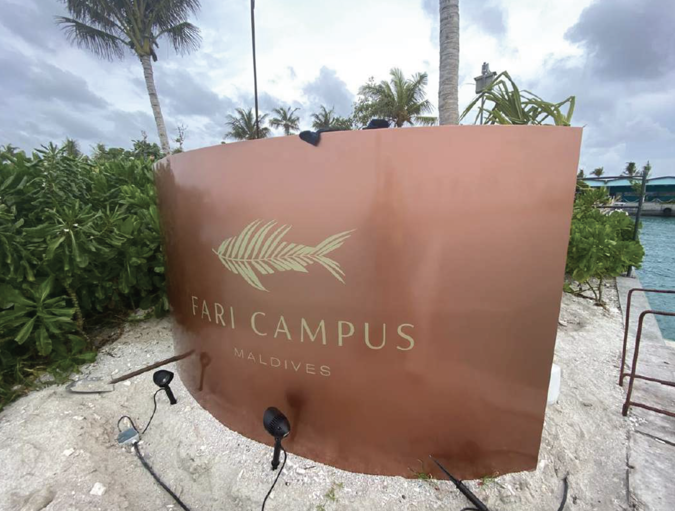 Sign for Fari Campus in Maldives, positioned outdoors near palm trees and water, with a rusty orange background and a white leaf graphic.