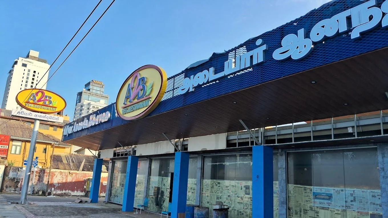 Exterior of a building with signs in multiple languages, including English and Sinhalese, advertising a restaurant and an educational institution, with high-rise buildings in the background, and a clear blue sky.