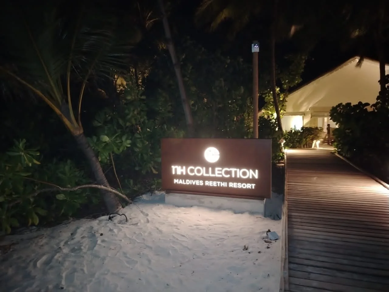 Illuminated sign at night reading 'TH COLLECTION MALDIVES REETHI RESORT' on sandy ground near a wooden walkway surrounded by tropical plants.