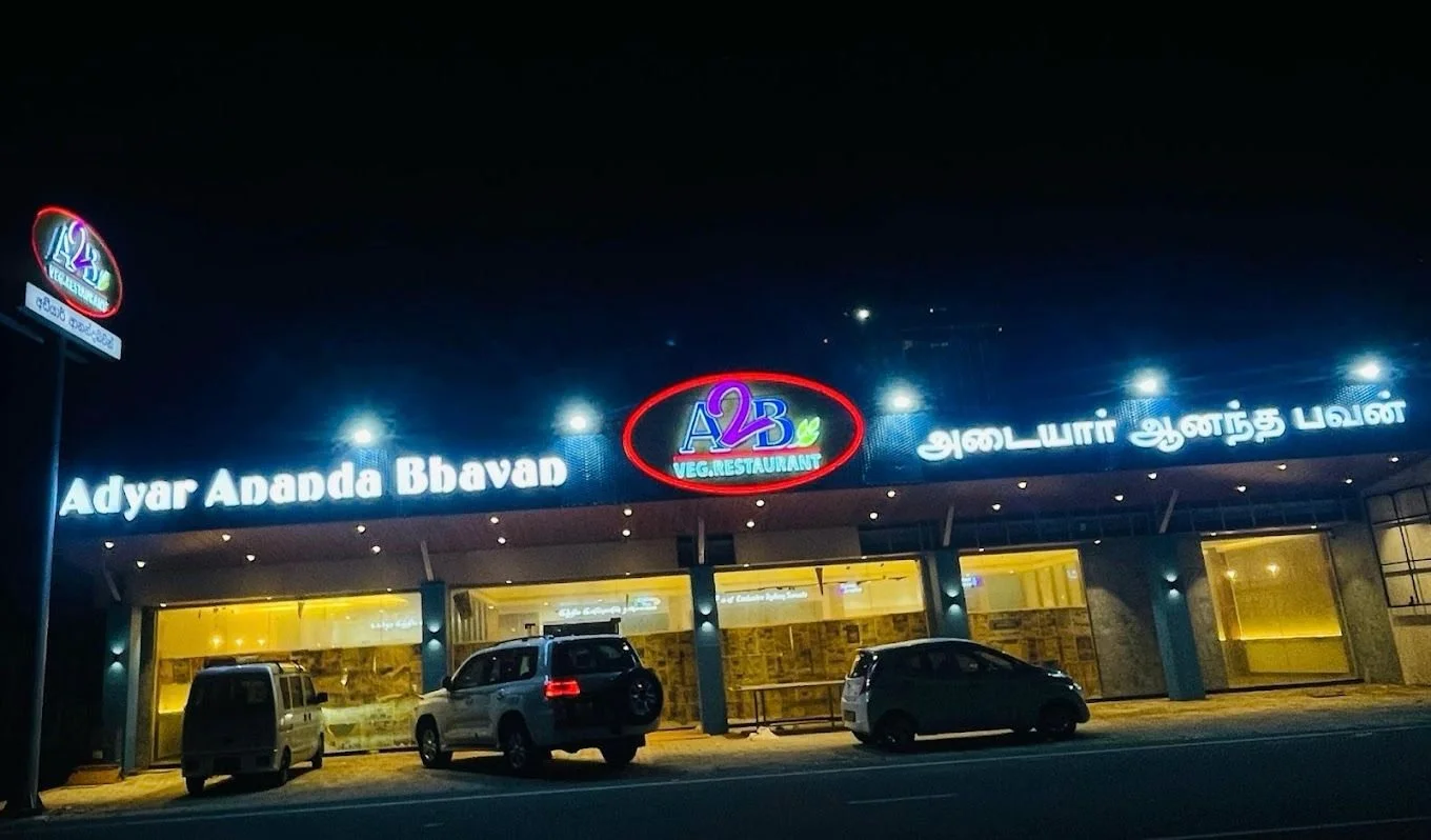 Night view of a restaurant with illuminated signage reading 'A2B Veg Restaurant' and 'Adyar Ananda Bhavan' in Tamil, with parked cars in front.