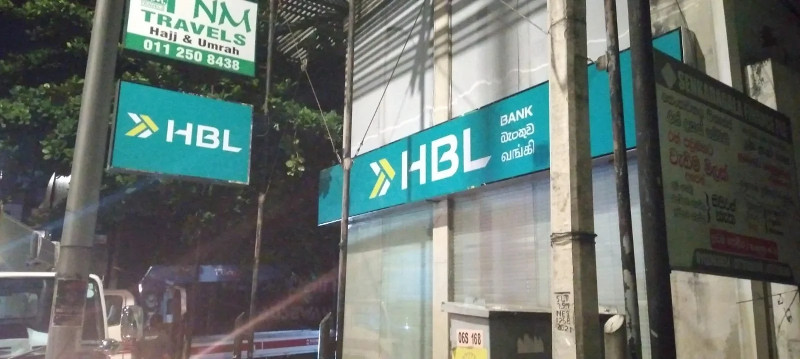 Night view of an HBL Bank sign on a building, with an additional HBL sign on a street pole, and a green sign for travel agency on top, all illuminated.