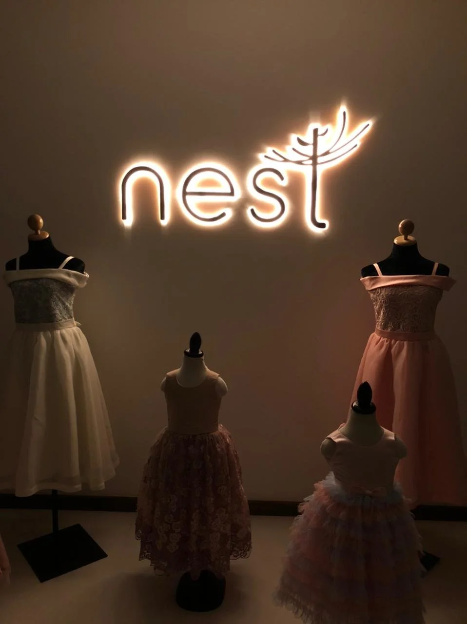 Neon sign spelling 'nest' with a bird's nest illustration, displayed above mannequins dressed in children's dresses at a boutique or clothing store.