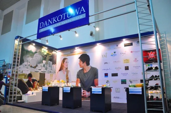 Trade show booth with a blue DANKOTUWA sign, displaying a large image of a man and woman talking, and shelves with shoes to the right.