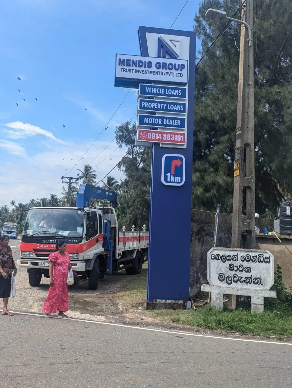 Signboard for Mendis Group Trust Investments indicating services like vehicle loans, property loans, and a motor dealer, with a contact number and a directional arrow showing 1 km ahead. There are two women walking by and a truck parked nearby, with 