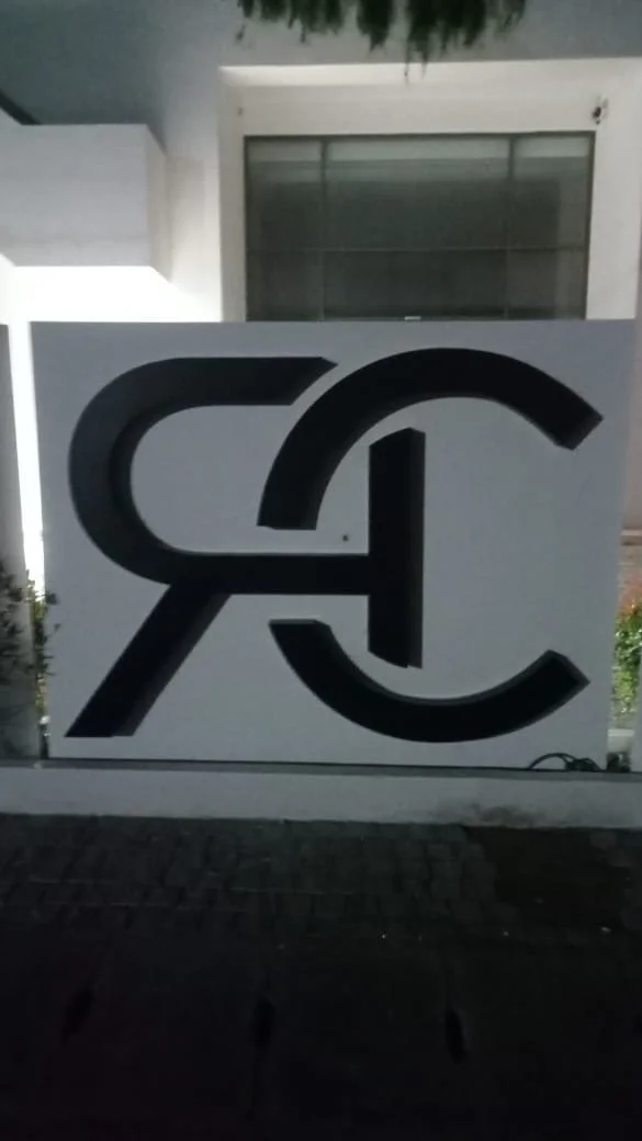 A large sign with stylized black letters: R and C, in front of a white building with a window.
