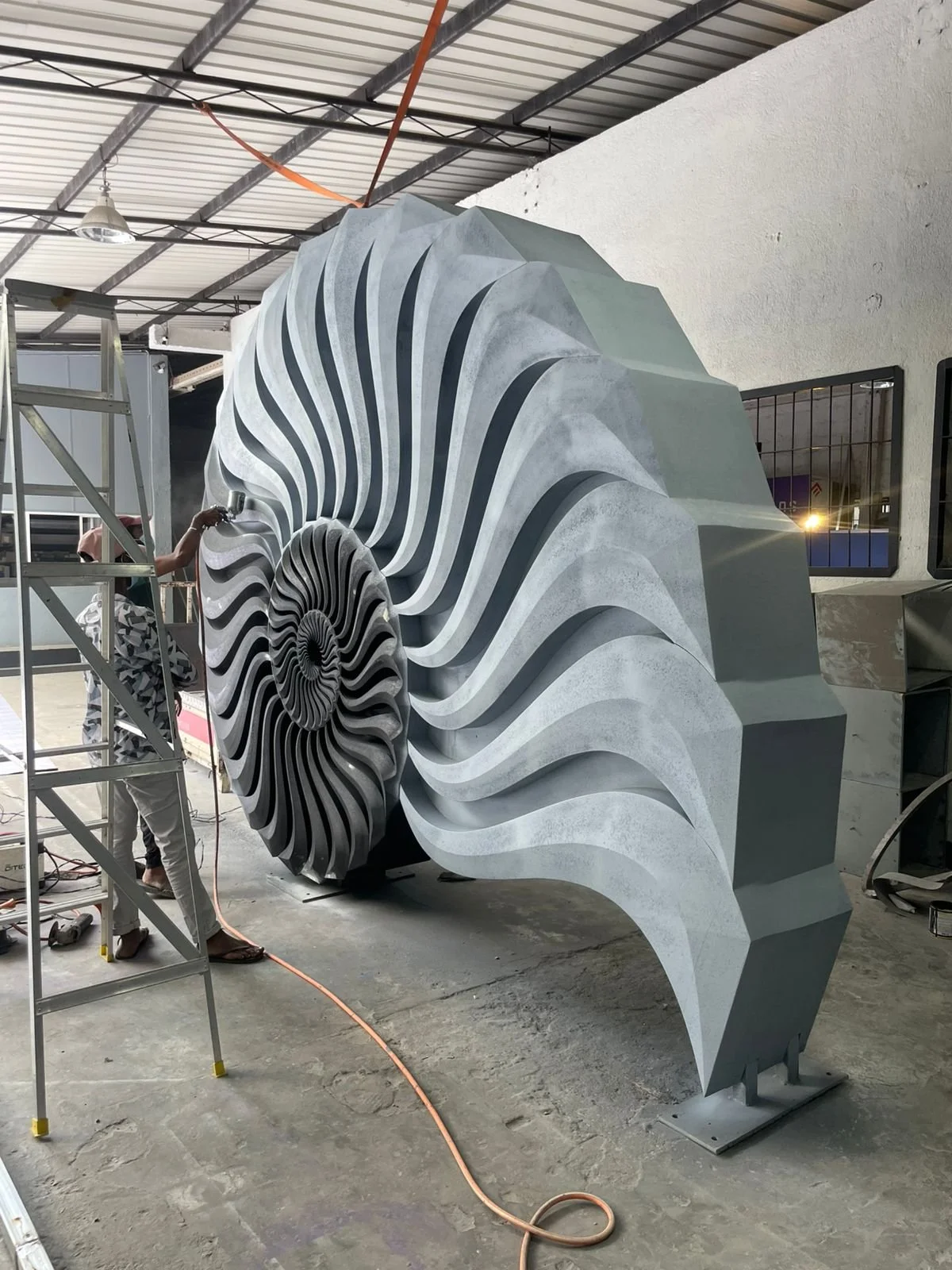 Workers are installing or constructing a large, modern, spiraling sculpture made of metal or concrete with a tunnel-like interior. The sculpture features a series of curved, flowing, layered ridges that create a spiral pattern.