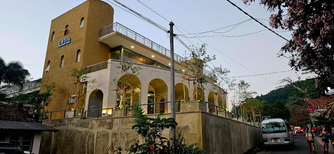 A multi-story building with a tan-colored upper section and white arches on the lower level, situated along a street with cars and pedestrians, surrounded by trees and power lines.