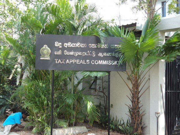 A sign for the Tax Appeals Commission in front of lush green plants and trees with a person crouching nearby.