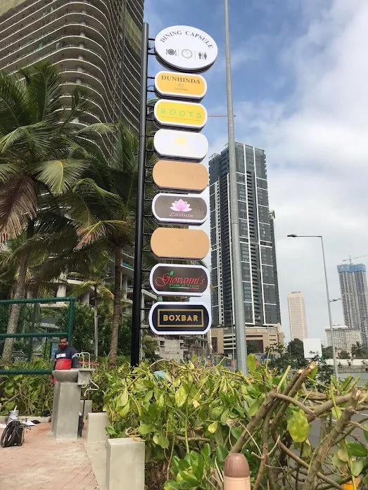 A tall signpost with multiple business signs, including a restaurant, a cafe, and a bar, with skyscrapers and a cloudy sky in the background.